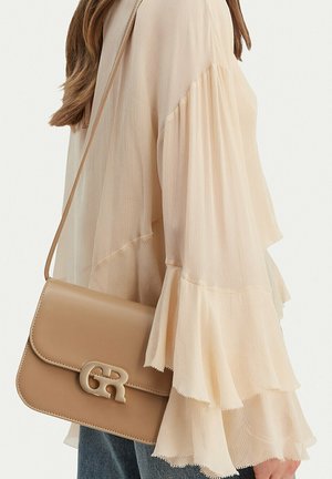 Mujer con una blusa beige de manga larga con volantes y un bolso de cuero marrón claro con un broche dorado decorativo.