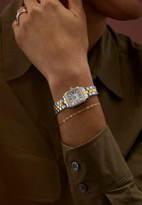 Silver- och guldfärgad metallarmbandsur med kvadratisk urtavla och urtavla med romerska siffror, visad på en hand som bär ett fint guldarmband.