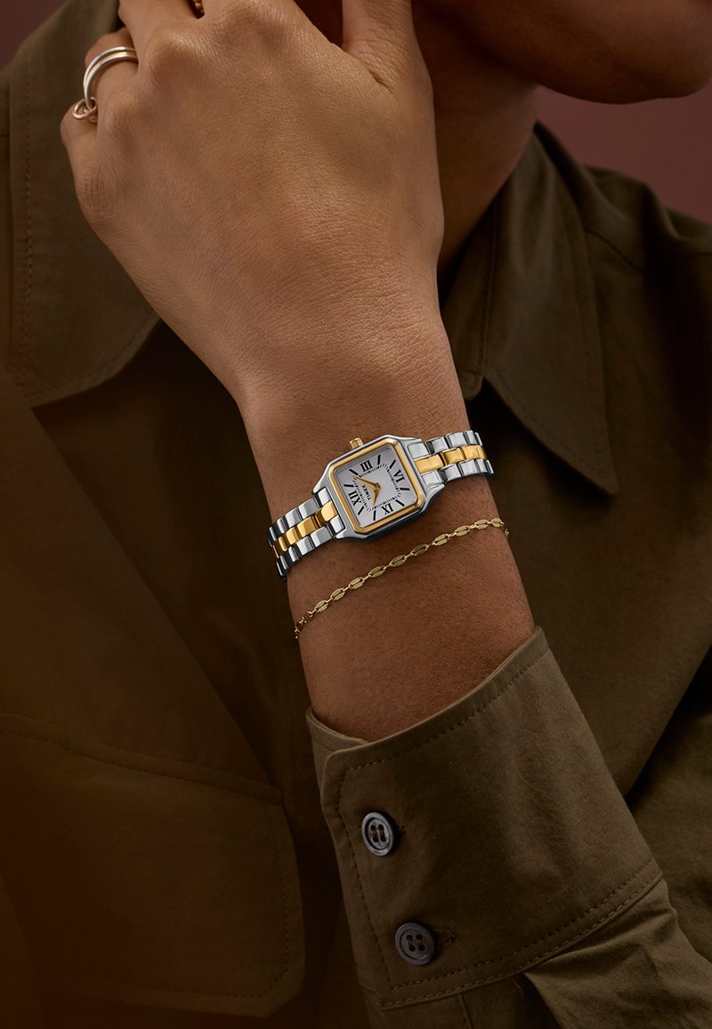 Silver- och guldfärgad metallarmbandsur med kvadratisk urtavla och urtavla med romerska siffror, visad på en hand som bär ett fint guldarmband.