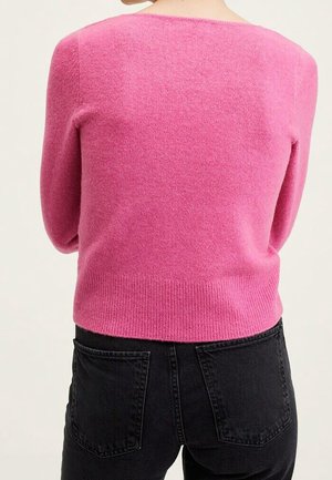 Mujer con un suéter de manga larga de color rosa brillante y jeans negros de talle alto, vista desde atrás contra un fondo liso.