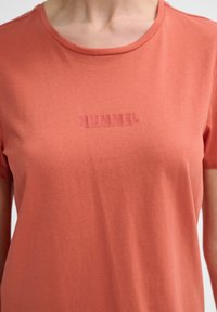 Koralfarvet bomulds t-shirt med rund halsudskæring, der har en blød tekstur og et præget "HUMMEL" logo på brystet. Korte ærmer.