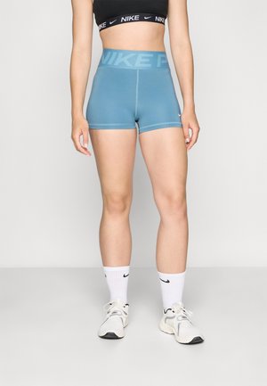 Femme portant un soutien-gorge de sport Nike noir, un short Nike Pro bleu taille haute, des chaussettes Nike blanches et des baskets blanches, debout devant un fond uni.