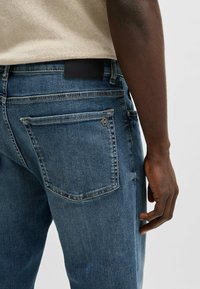 Jeans de mezclilla azul oscuro con un corte recto. Presentan un parche de cuero negro de marca y detalle metálico en el bolsillo trasero.