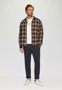 Mannenoutfit met een zwart-en-bruin geblokt flanellen shirt, een witte crewneck t-shirt, donkerblauwe broek en witte sneakers.