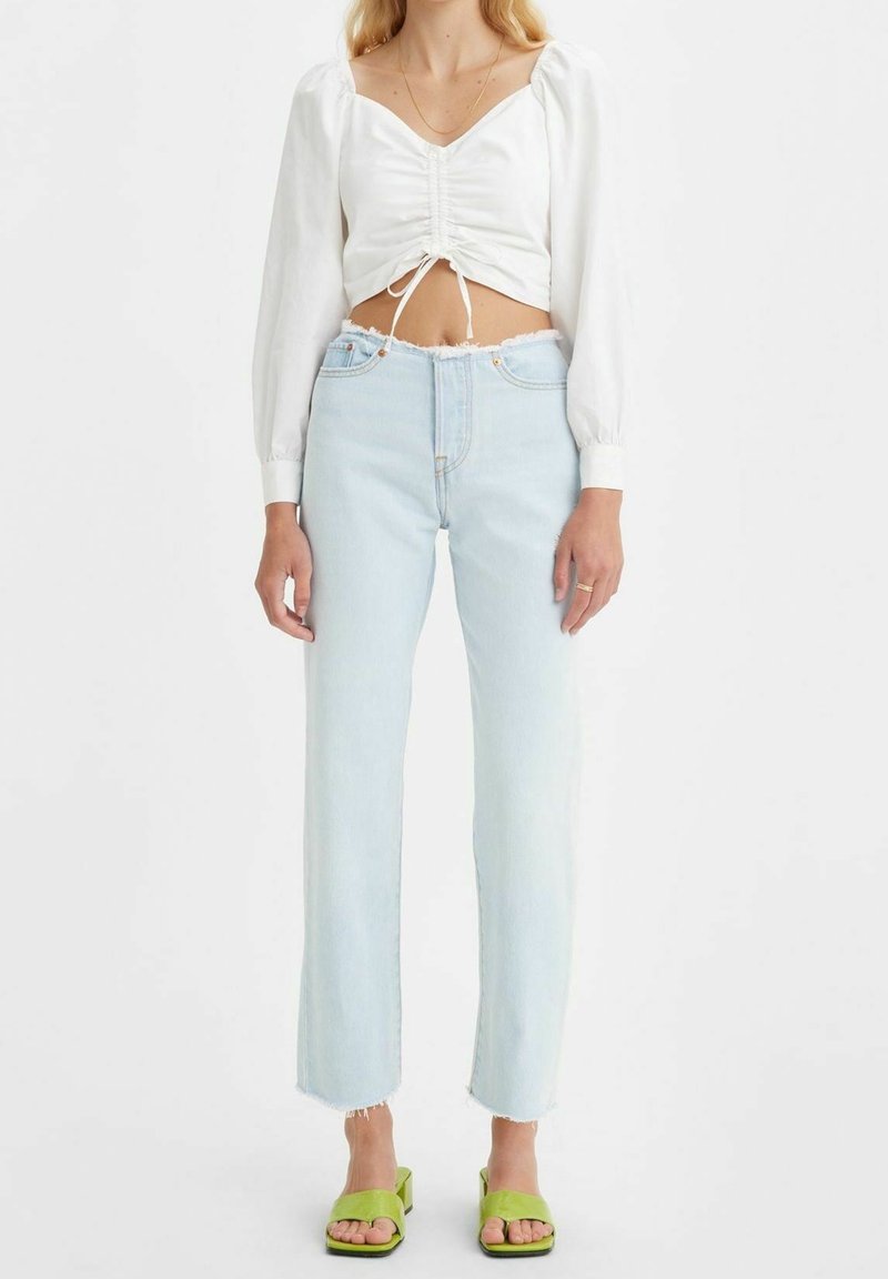 Haut court à manches longues blanc avec un détail froncé, associé à un jean taille haute bleu clair ; des sandales à talons blocs vertes complètent la tenue.