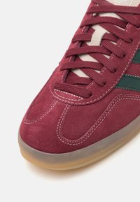 Burgunderfargede semskede sneakers med beige snørebånd, gummisåle og grønne striper langs siden. Har buet søm og teksturert overflate.