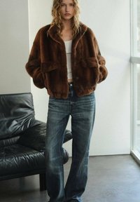 Chaqueta de piel sintética marrón con un corte relajado, cierre de botones y dos bolsillos frontales, combinada con unos pantalones vaqueros de pierna ancha de lavado claro.