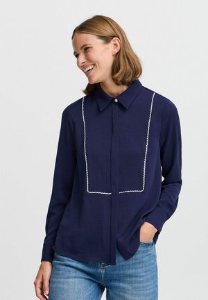 Navy blouse with a button-up front, collar, and white contrasting trim outlining a rectangular design on the chest, paired with denim jeans.