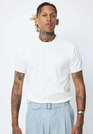 T-shirt blanc à manches courtes avec un col rond, texture douce, porté avec un pantalon plissé bleu clair et une montre en argent.
