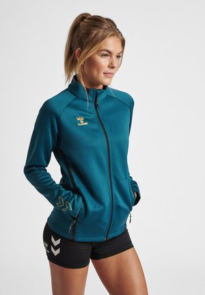 Kobieta z blond włosami związanymi w kucyk, ubrana w turkusową kurtkę zapinaną na zamek i czarne sportowe szorty, ręce w kieszeniach kurtki, zwrócona w prawo.