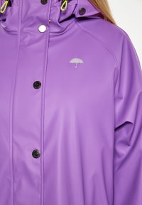 Giacca impermeabile viola con una texture liscia, bottoni a pressione e un piccolo logo di un ombrello argentato sul petto. Cappuccio regolabile con dettagli gialli.