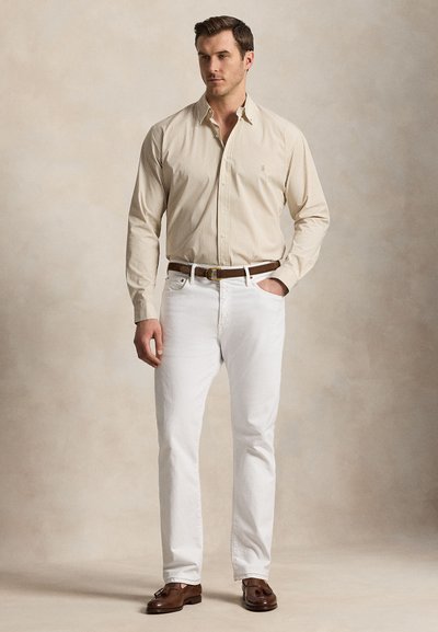 Man draagt een beige gestreept overhemd met lange mouwen en knopen, witte broek, bruine riem en bruine loafers met kwastjes, staat met één hand in de zak.