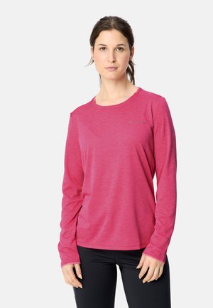 Frau mit braunen Haaren im Pferdeschwanz, die ein langärmeliges pinkes Shirt und schwarze Hose trägt und vor einem einfarbig hellen Hintergrund steht.