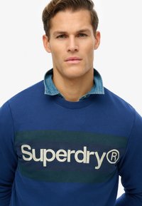 Niebieska bluza z ciemnozielonym poziomym paskiem i białym logo "Superdry". Kołnierz jest dżinsowy, co dodaje kontrastu i tekstury.