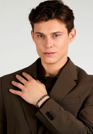 Giacca da suit marrone su misura con bottoni neri, indossata sopra una camicia scura. Un bracciale argento con motivi geometrici è visibile sul polso sinistro.