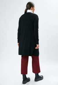 Lange zwarte cardigan met een rechte snit, gedragen over bordeauxrode broek en zwarte enkellaarsjes. Heeft ribbelmanchetten en een ribbelzoom.