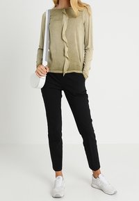 Maglione lungo verde oliva con un dettaglio arricciato centrale, abbinato a pantaloni neri slim-fit e sneakers bianche, con una piccola borsa a tracolla bianca.