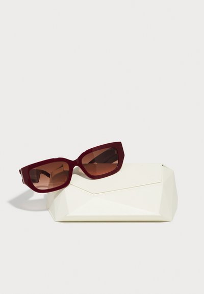 Red oversized sunglasses with a geometric frame and gradient brown lenses, placed on a cream, angular protective case.