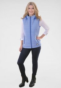 Hellblau gefütterte Weste mit hohem Kragen, Reißverschluss und Seitentaschen, getragen über einem weißen Langarmshirt. Schwarze Knöchelstiefel.