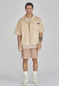 Camicia beige a maniche corte con bottoni e due tasche frontali, abbinata a una maglietta beige chiaro e pantaloni cargo color khaki. Calzini bianchi e sneakers.