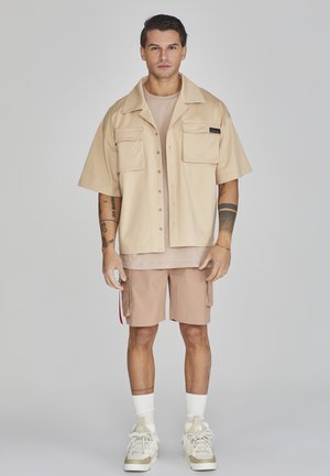 Beige Kurzarmhemd mit Knöpfen und zwei vorderen Taschen, kombiniert mit einem hellbeigen T-Shirt und khakifarbenen Cargo-Shorts. Weiße Socken und Sneaker.