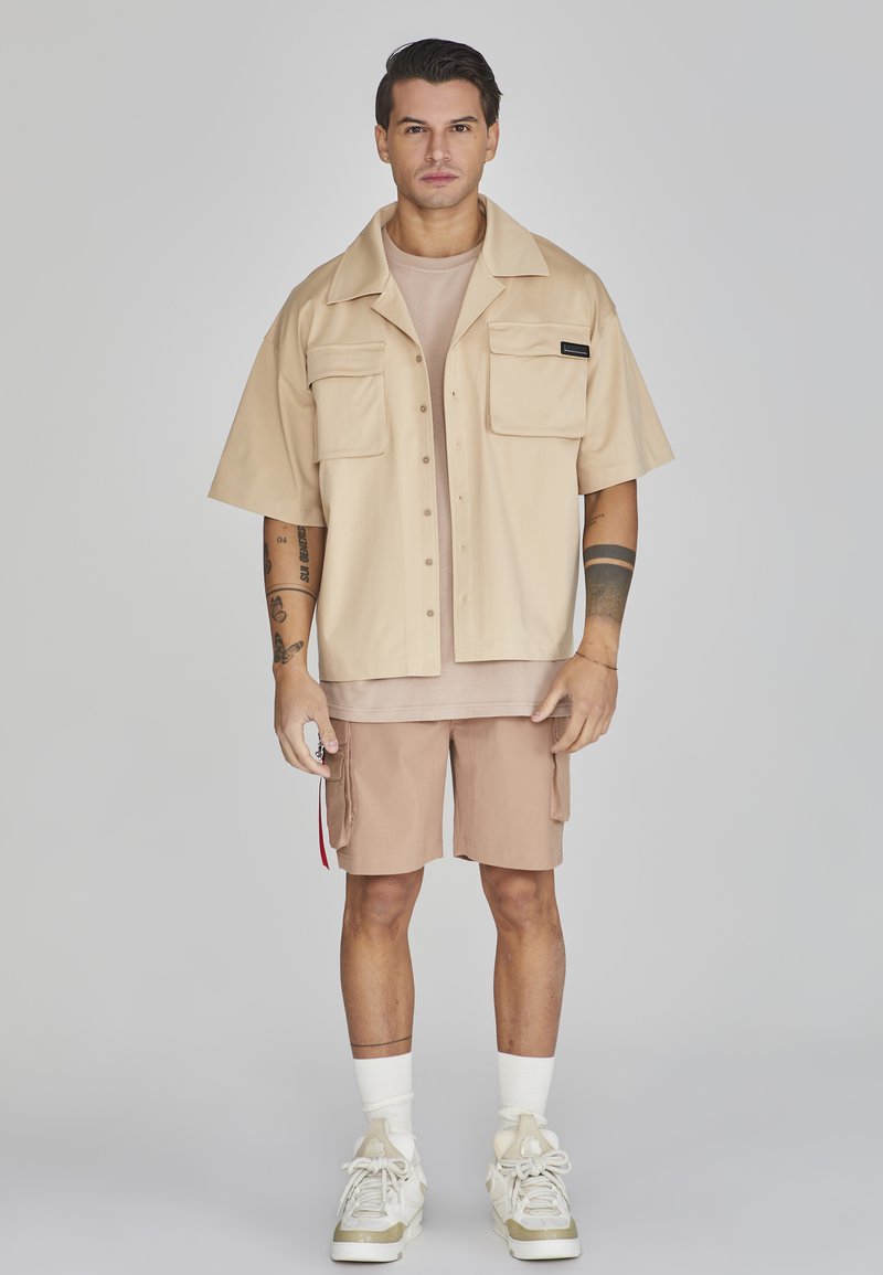 Camicia beige a maniche corte con bottoni e due tasche frontali, abbinata a una maglietta beige chiaro e pantaloni cargo color khaki. Calzini bianchi e sneakers.