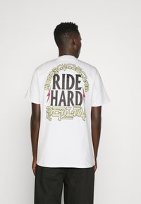 Camiseta de algodón blanca con un gran gráfico en la parte trasera, letras en negrita, acentos de relámpagos y un diseño retro.