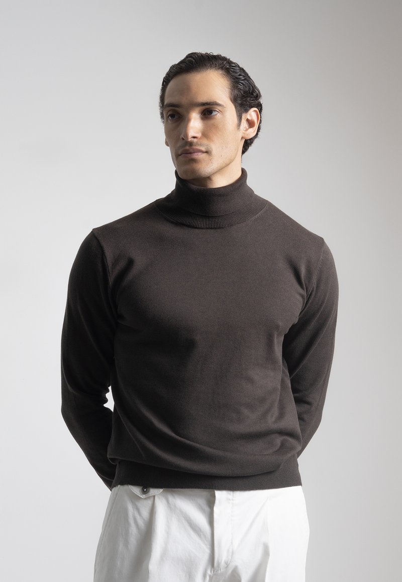 Hombre con cabello rizado oscuro, vistiendo un suéter de cuello alto marrón oscuro y pantalones blancos, de pie contra un fondo claro y simple.