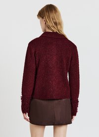 Bordeauxroter, strukturierter Blazer mit Kragen und Fransen an den Manschetten, kombiniert mit einem dunkelbraunen Minirock. Das Material wirkt weich und gewebt.