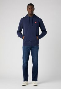 Granatowa bluza z kapturem z kieszenią kangurką z przodu oraz logo na piersi, w połączeniu z niebieskimi dżinsami i białymi trampkami. Gładka tekstura, casualowy styl.