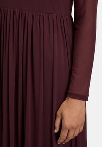 Bordeauxfarbenes Kleid mit langen, durchsichtigen Ärmeln. Mit einem plissierten Rock und einem strukturierten Oberteil. Glatter Stoff mit subtiler glänzender Oberfläche.