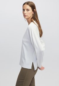 Witte pullover trui met een ontspannen pasvorm, voorzien van een ronde halslijn, zijsplitten en lange mouwen. Zachte textuur; gecombineerd met olijfgroene broek.