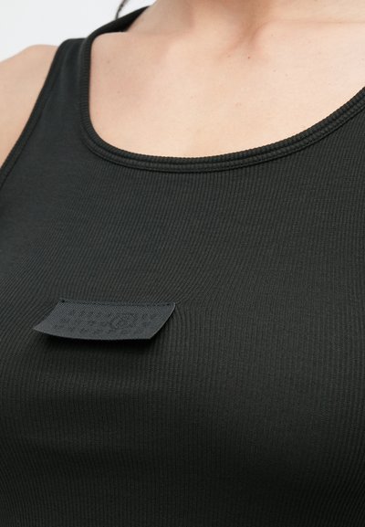 Svart ribbestrikket tanktopp med rund hals og en strukturert svart stofflapp sydd på forsiden, nærbilde.