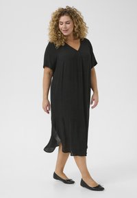 Robe noire ample avec un décolleté en V et des manches courtes, dotée d'un tissu texturé et de fentes sur les côtés pour plus de liberté de mouvement. Portée avec des ballerines noires.