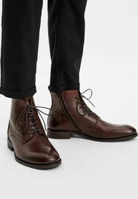 Bottines en cuir marron avec des lacets et détails de fermeture éclair sur le côté. Bout arrondi et talon bas, assorties à un pantalon noir ajusté.