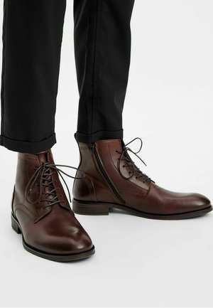 Botas de cuero marrón hasta el tobillo con detalles de cordones y cremallera lateral. Punta redonda y tacón bajo, combinadas con pantalones negros ajustados.