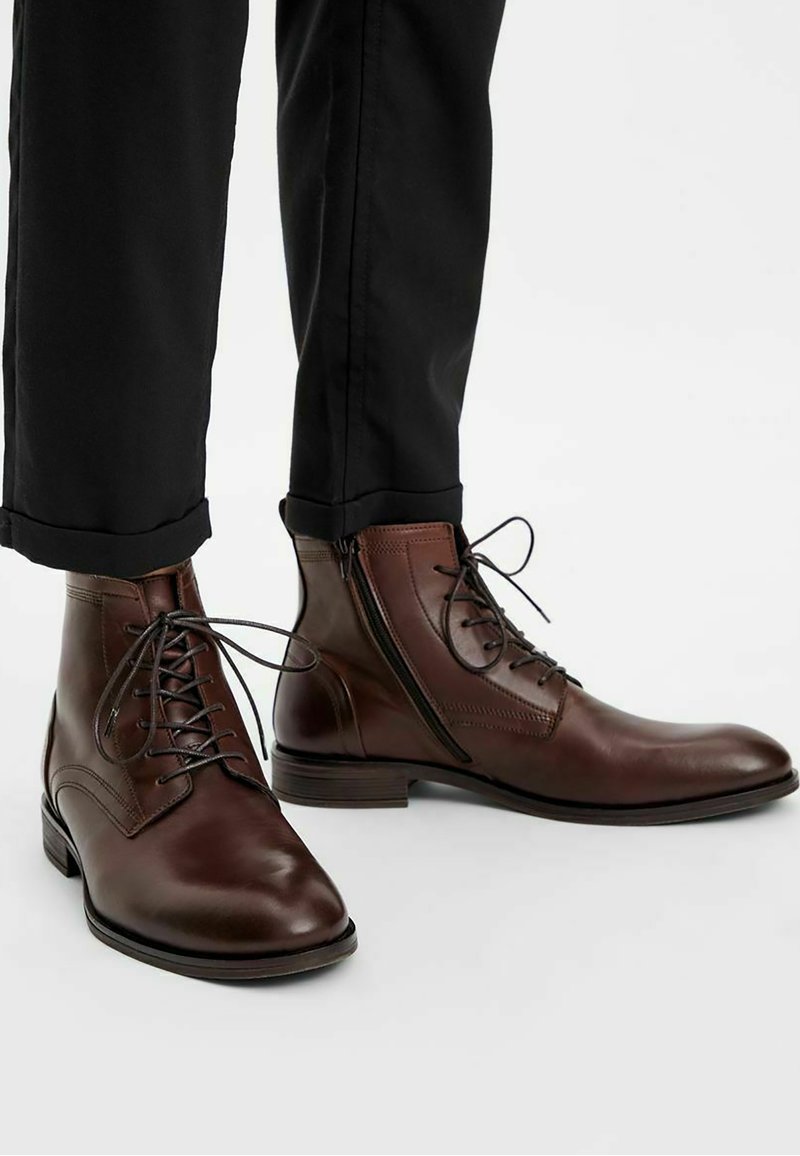 Bottines en cuir marron avec des lacets et détails de fermeture éclair sur le côté. Bout arrondi et talon bas, assorties à un pantalon noir ajusté.