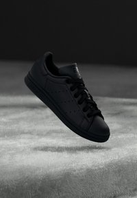 Černý teniska Adidas Stan Smith se šněrováním, vznášející se nad texturovaným šedým povrchem na tmavém pozadí.