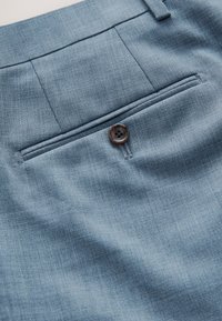 Poche arrière d’un pantalon de costume bleu clair avec un seul bouton marron et une boucle de ceinture visible à la taille.