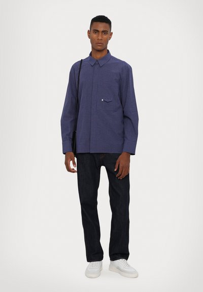 Chemise à carreaux bleu marine avec une poche avant, associée à un jeans en denim foncé et des baskets blanches. Coupe décontractée avec des manches longues et un col.