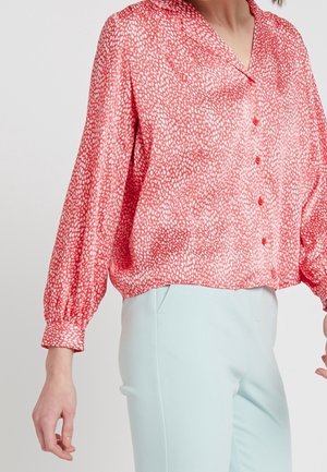 Woman wearing a red patterned long-sleeve button-up blouse with light blue pants, standing against a plain background.