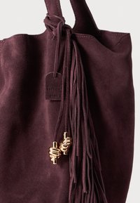 Burgundy suede tote bag with long fringe detailing, gold hardware accents, and a tag reading "Genuine Leather" attached.