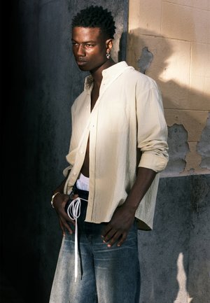 Chemise boutonnée texture jaune clair portée ouverte sur une ceinture blanche ; pantalon en denim bleu à jambes larges avec cordon de serrage ; manches courtes.