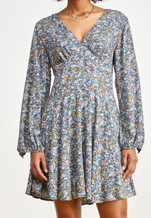 Femme portant une robe à manches longues, à encolure en V, longueur genou, avec un motif floral bleu, blanc et orange et une taille froncée.