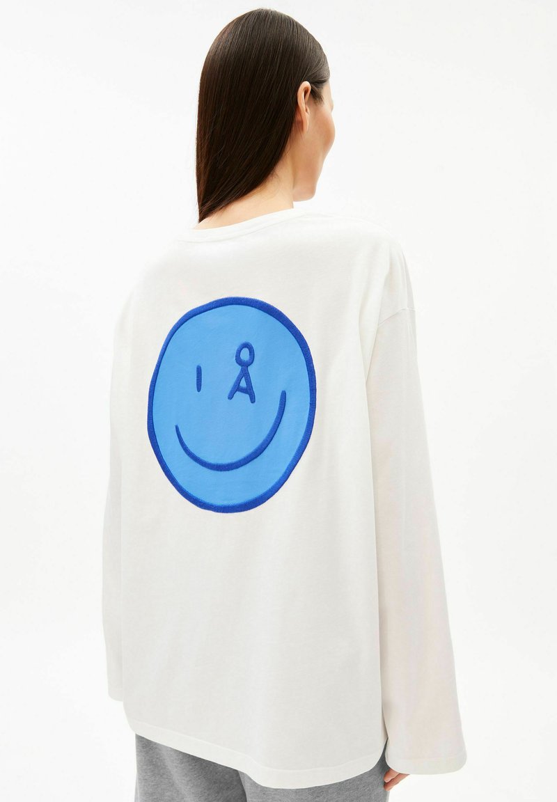 Femme aux longs cheveux foncés portant une chemise blanche à manches longues avec un grand graphique de visage souriant bleu dans le dos, sur un fond blanc uni.