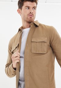 Beige Hemd mit Knöpfen und einer Brusttasche, aus leichtem Material gefertigt. Verfügt über einen Kragen und einen lockeren Schnitt, über einem weißen T-Shirt getragen.