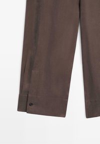 Braune gerade geschnittene Hose mit Knopfdetail am Saum vor hellem Hintergrund.