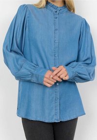 Camicia in denim azzurro chiaro con colletto a volants, chiusura con bottoni sul davanti e maniche lunghe a sbuffo. Il tessuto appare liscio e ha una vestibilità comoda.