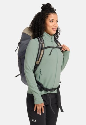Donna con capelli ricci che indossa una giacca verde con zip e pantaloni neri, che porta uno zaino da trekking grande grigio e beige.