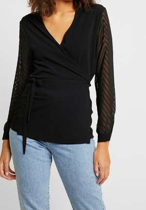 Femme portant un top cache-cœur noir avec des manches longues transparentes et à motifs, et un jean bleu clair, debout devant un fond clair uni.